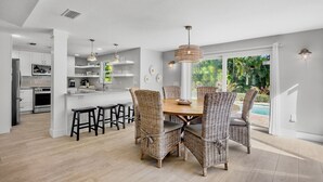 A modern kitchen with white cabinets, a central island, and a dining area with wicker chairs and a wooden table. - C-View Florida: Canal Front Home w/ Dock! (Anna Maria)