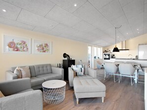Een moderne woonkamer met een bank, poef en een rond salontafeltje, een eethoek met stoelen, en een keuken met een fornuis en spoelbak.