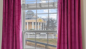 Ein Fenster mit rosa Vorhängen, Blick auf ein Gebäude mit einem gelben Schild, und ein weißer Zaun im Hintergrund.