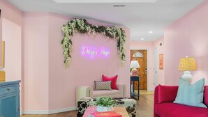 Ein pinkfarbenes Wohnzimmer mit einem Neon-Schild mit der Aufschrift „Dolly's House“, einem Sofa, einem Couchtisch mit Büchern und einer Zierpflanze.