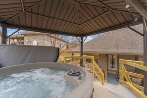 A hot tub under a covered patio with a wooden railing, a brick house in the background, and a clear sky. - Dolly's House! In the heart of Branson! (Branson)