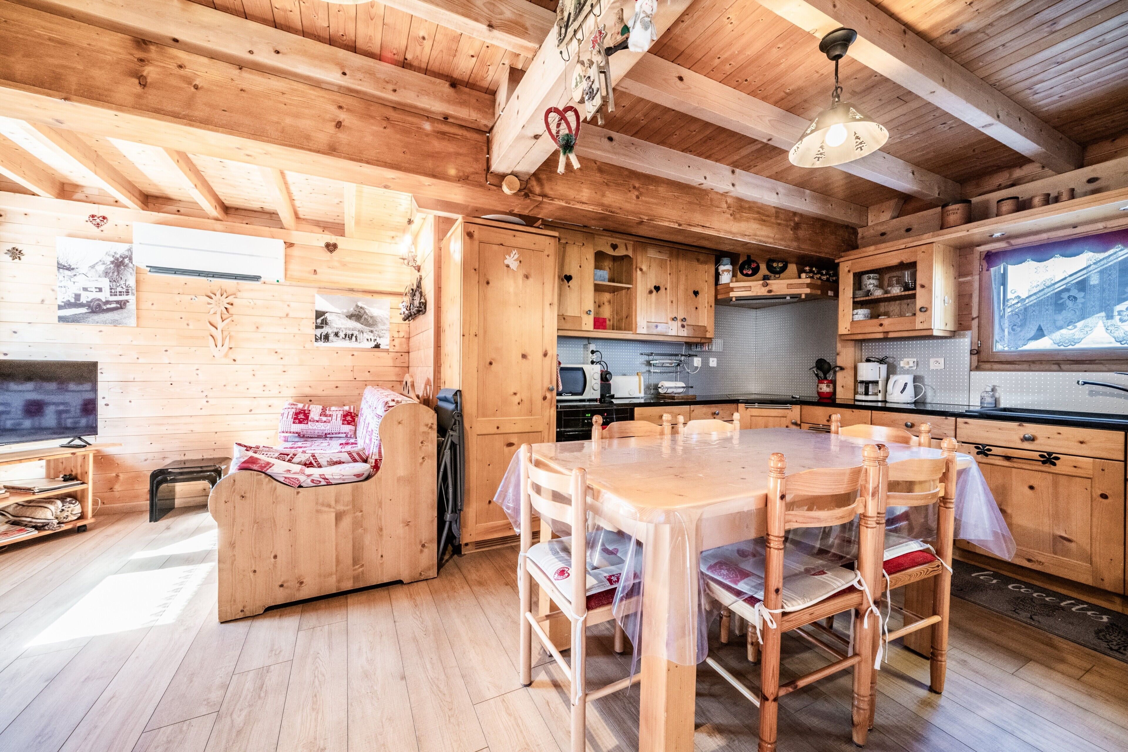 Interno di una baita in legno con tavolo da pranzo, sedie, divano, angolo cottura e televisore.