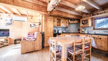 Interno di una baita in legno con tavolo da pranzo, sedie, divano, angolo cottura e televisore.