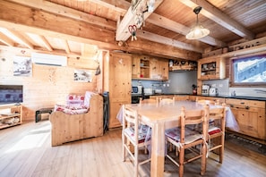 A wooden cabin interior with a dining table, chairs, a sofa, kitchen area, and a television. - Chalet “6p Premium - La Bergeronette” with Mountain View, Wi-Fi, and Air Conditioning (Verchaix)