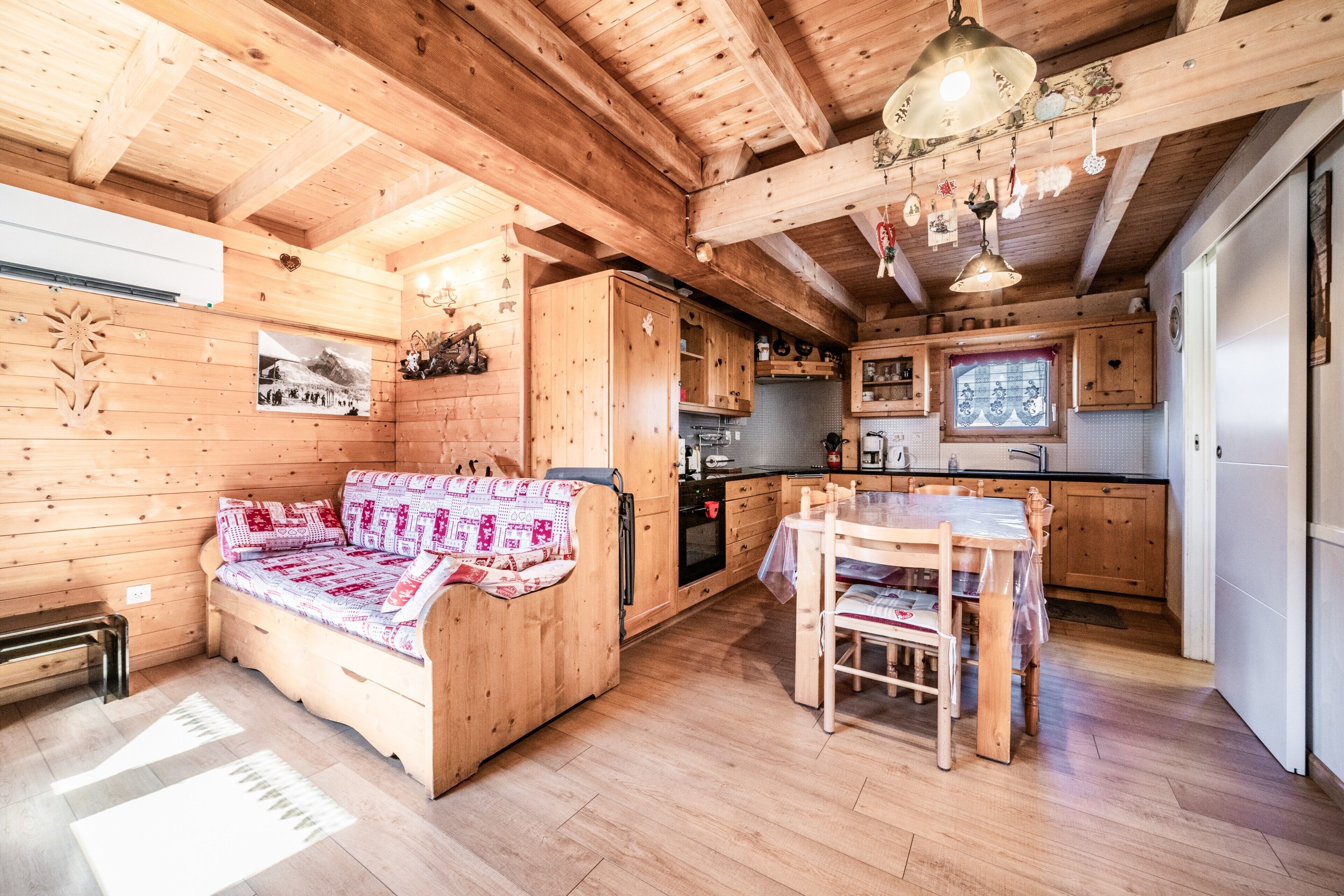 Interno di una baita in legno con divano, tavolo da pranzo, angolo cottura e travi a vista al soffitto.