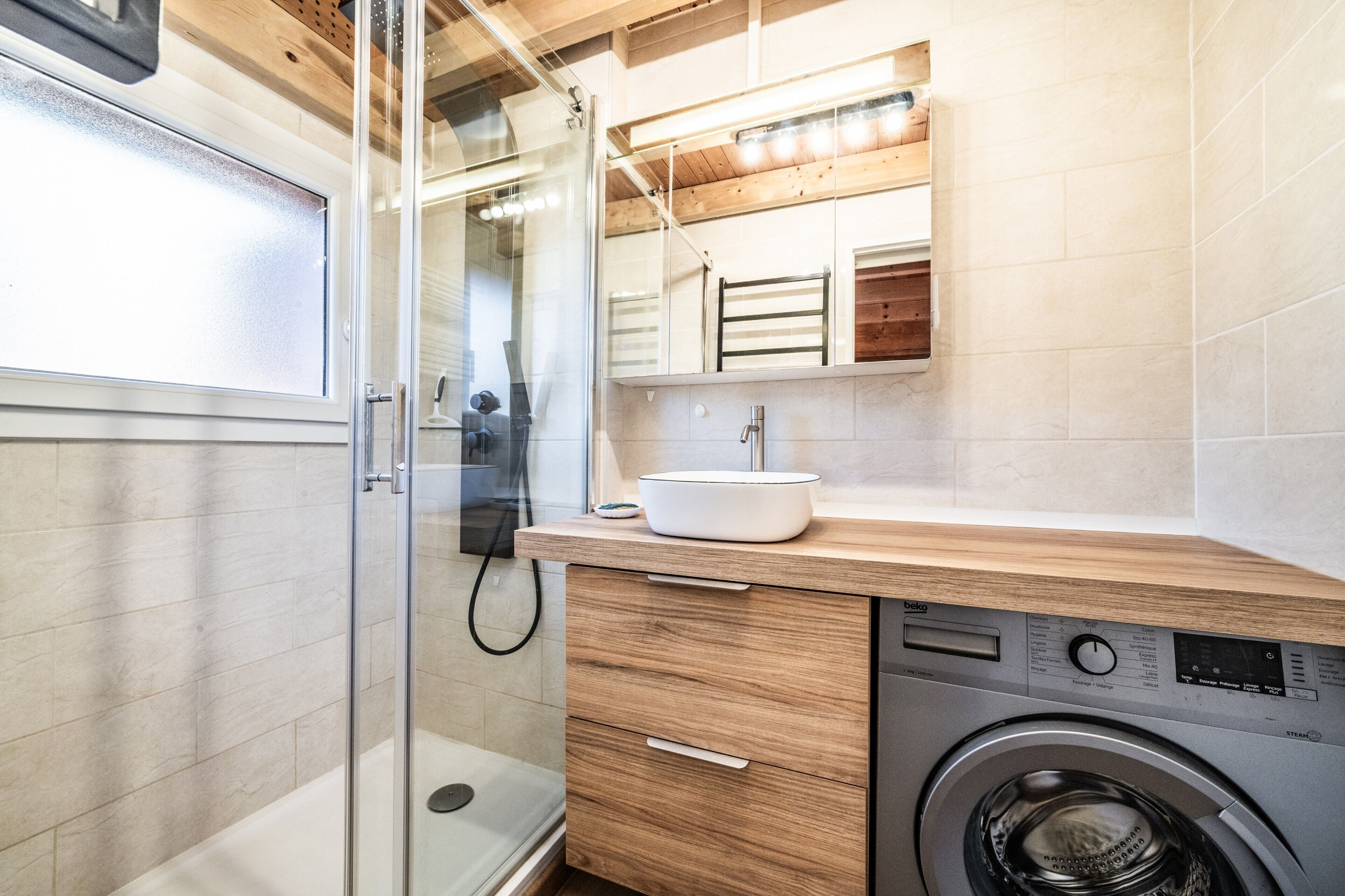 Un baño moderno con cabina de ducha de vidrio, lavadora integrada y tocador de madera con lavamanos.