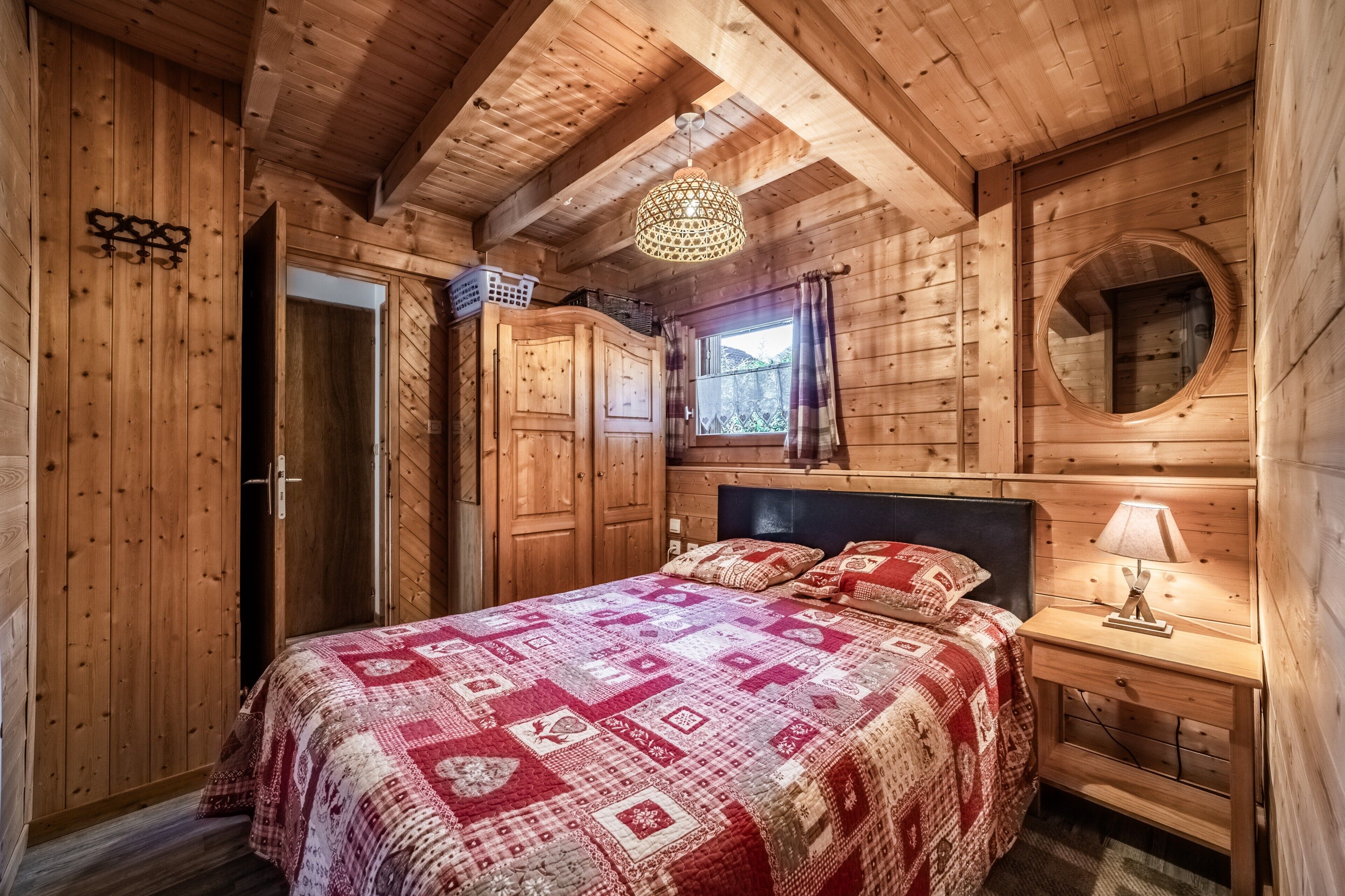 A wooden cabin bedroom with a bed, a nightstand, a hanging light, a wardrobe, and a circular window.