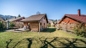 Une maison en bois avec un toit en pente, une terrasse avec une table et des chaises, et une cour gazonnée.