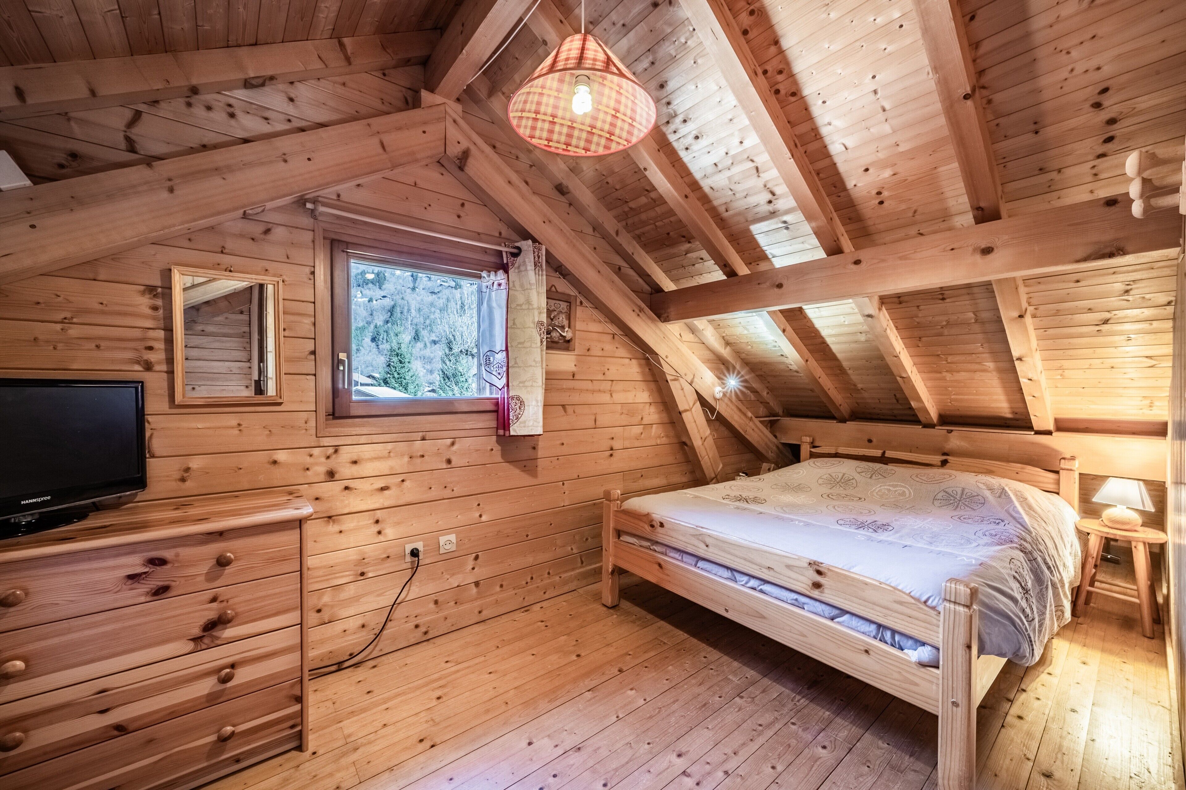 Camera da letto in una baita di legno, con un letto, una cassettiera, una TV e una finestra vista alberi.
