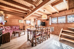 Een houten blokhut van binnen met een eettafel, stoelen, een bank en een keukenhoek.