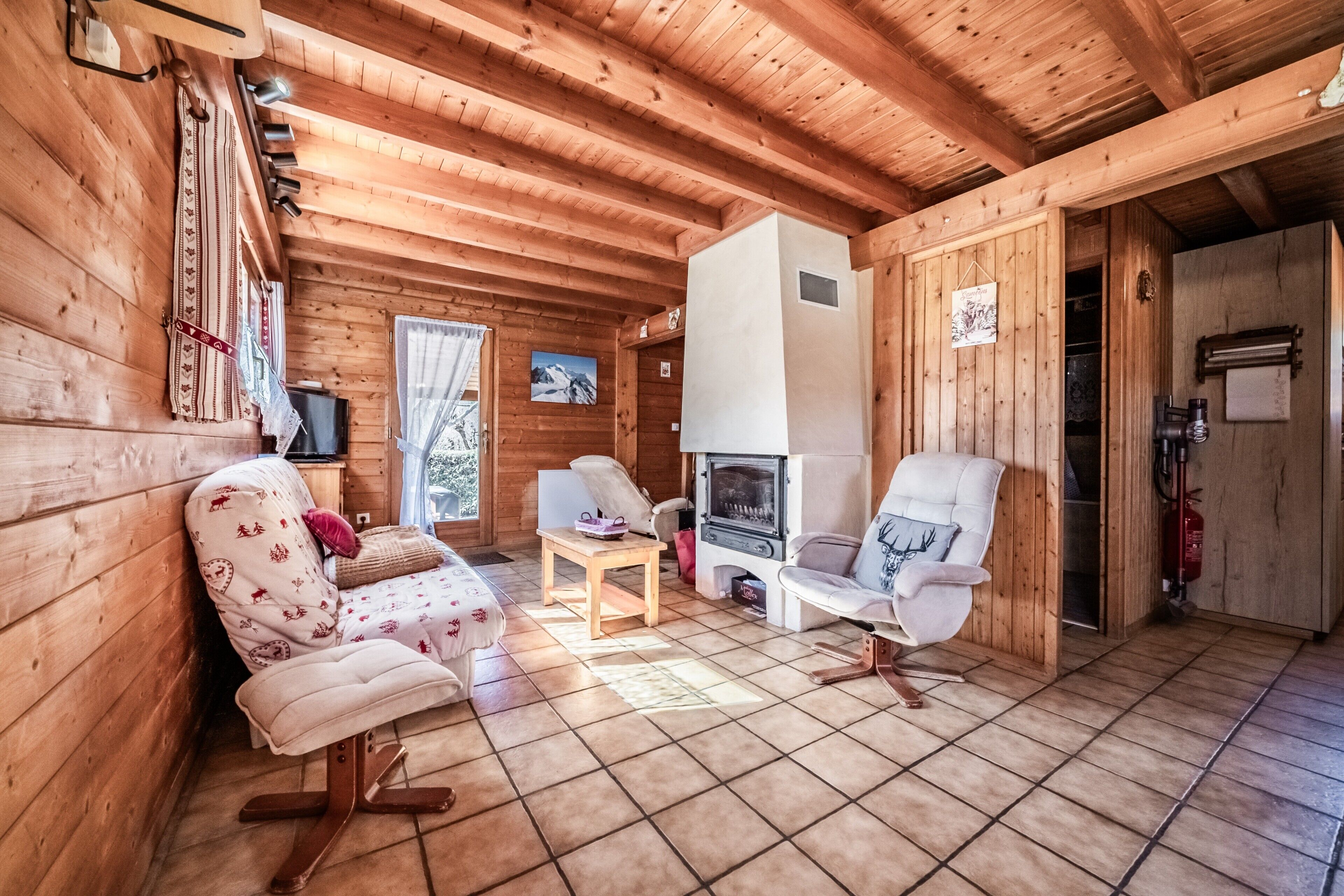 Interior de una cabaña de madera con sofá, dos sillones, una mesita, chimenea y ventana con vistas a montañas nevadas.