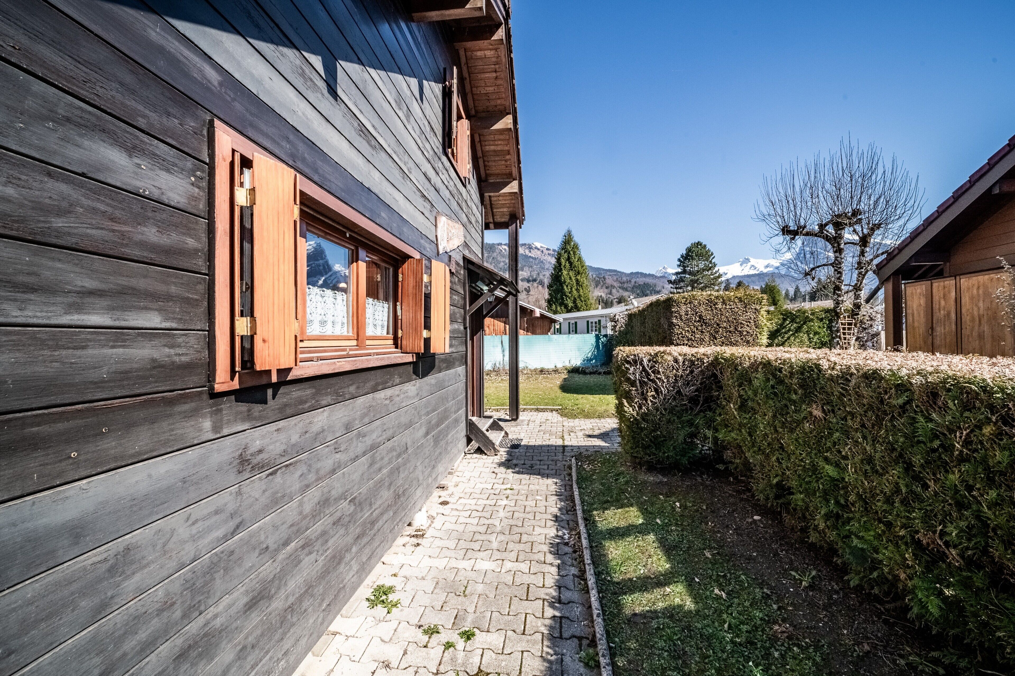 Un edificio in legno con un vialetto lastricato che conduce a una zona verde, una recinzione in legno e una vista chiara su una catena montuosa.