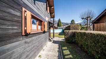 Un edificio in legno con un vialetto lastricato che conduce a una zona verde, una recinzione in legno e una vista chiara su una catena montuosa.