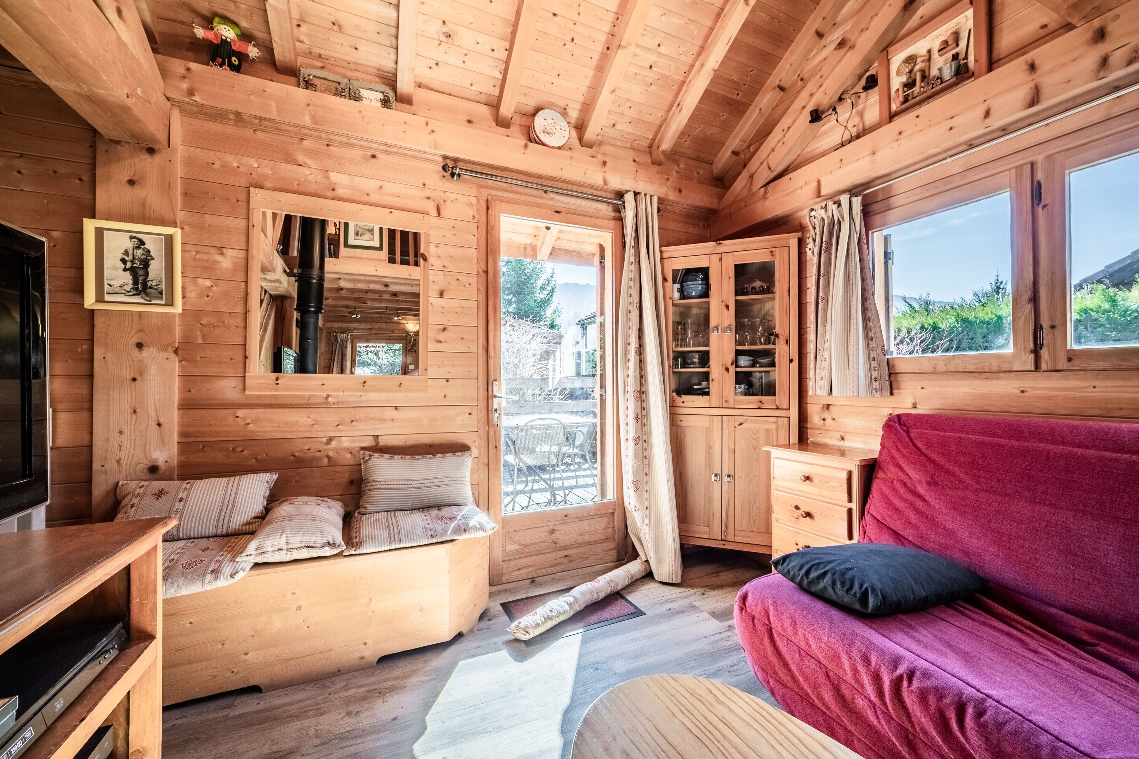 Interior de una cabaña de madera con sofá, televisión, zona de comedor y ventana con vistas.