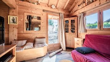 Interior de una cabaña de madera con sofá, televisión, zona de comedor y ventana con vistas.