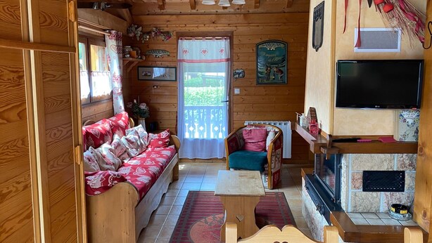 Un accogliente interno di una baita con pavimento in legno, un divano, una poltrona, un tavolo, un camino e un televisore.