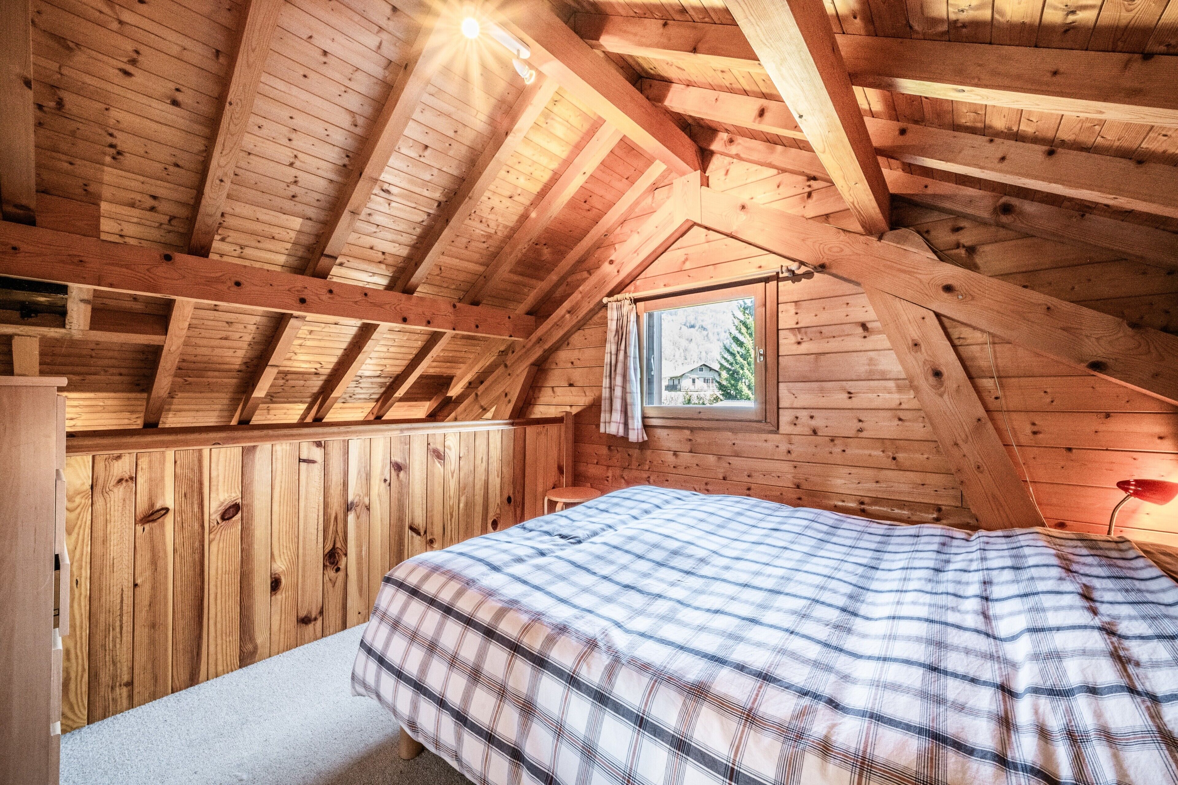Ein Schlafzimmer im Dachgeschoss mit Holzboden, einem Bett, einem Fenster mit Vorhängen und einer roten Lampe.