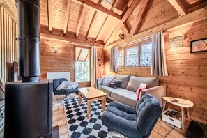 Un interior de cabaña acogedor, con un estufa de leña, un sofá, un sillón, una mesita y una ventana con cortinas.