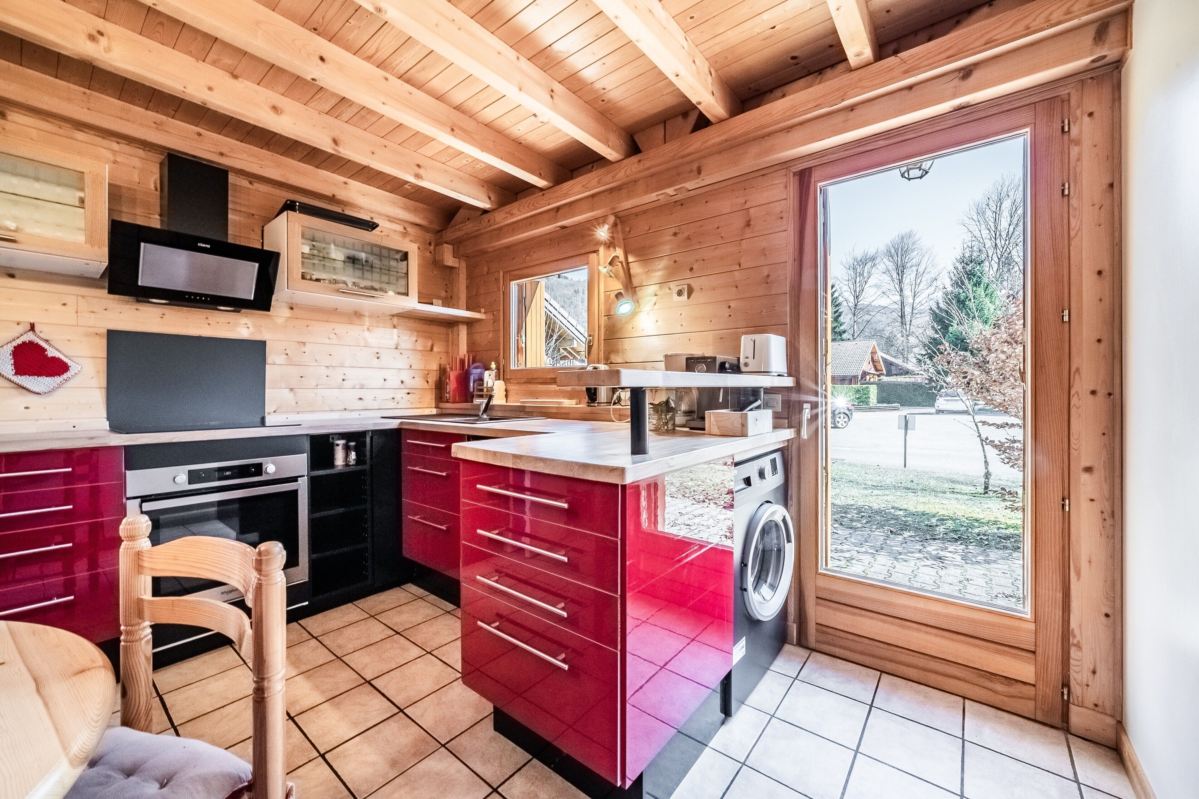 Interno di una baita di legno con cucina, zona pranzo e un'ampia porta-finestra che si apre verso l'esterno.