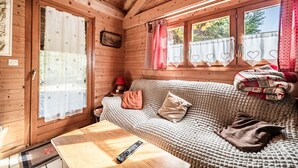 Ein gemütliches Blockhaus-Interieur mit Holzbalkendecke, einer Couch mit Decke, einer Fernbedienung auf einem Tisch und Vorhängen mit Herzmuster.