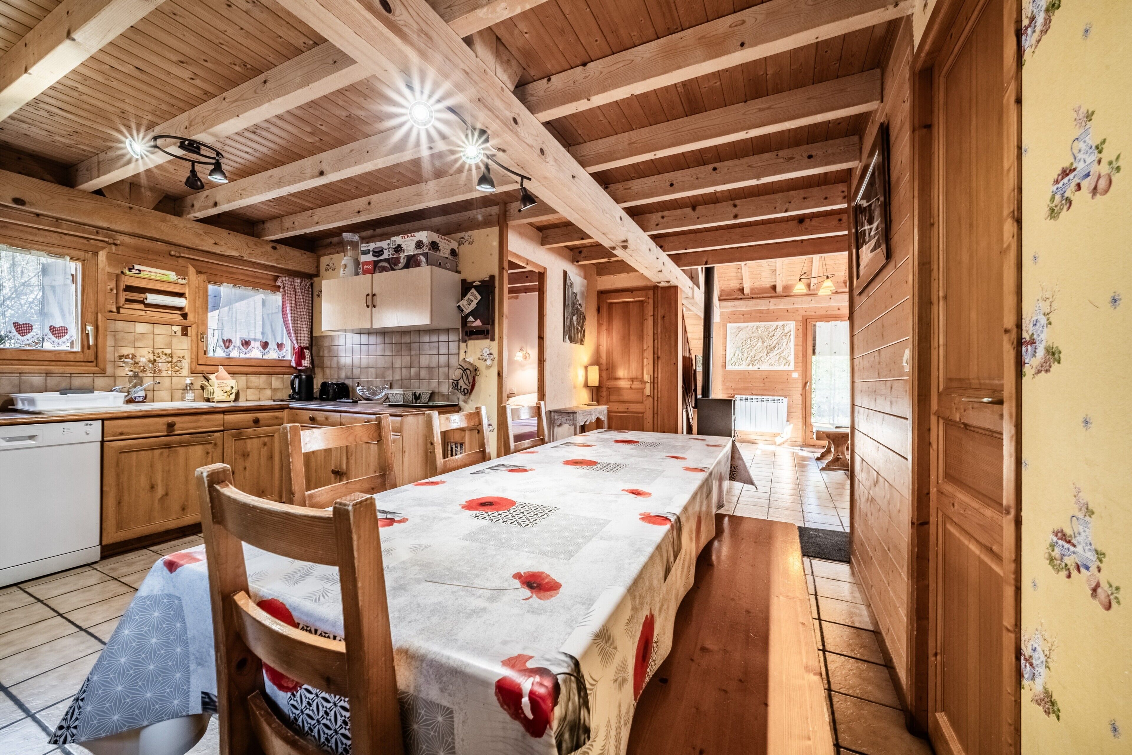 Interior de una cabaña de madera con mesa de comedor, zona de cocina y vigas de madera en el techo.