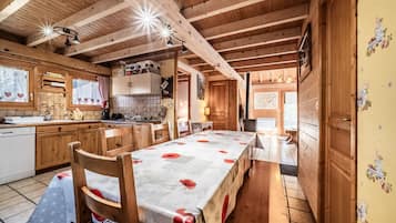 A wooden cabin interior with a dining table, kitchen area, and wooden beams on the ceiling.