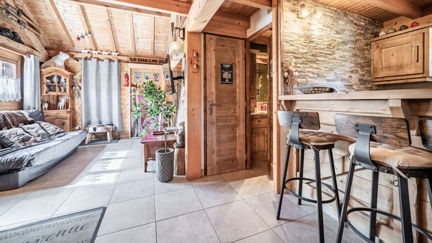 Et træinteriør med en sofa, et spiseområde med barstole, et køkken med stenvægge, og en dør med teksten 'Le Chalet 36'.