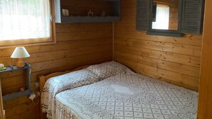A wooden cabin room with a bed, a window with lace curtains, a shelf with items, and a mirror with shutters. - Chalet “6p Comfort - Le Chevreuil”: Mountain View, Private Terrace & Wi-Fi (Verchaix)