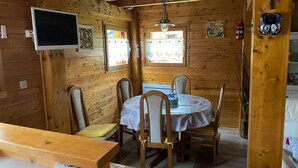A wooden cabin interior with a dining table, chairs, a TV mounted on the wall, and a window with a view. - Chalet “6p Comfort - Le Chevreuil”: Mountain View, Private Terrace & Wi-Fi (Verchaix)