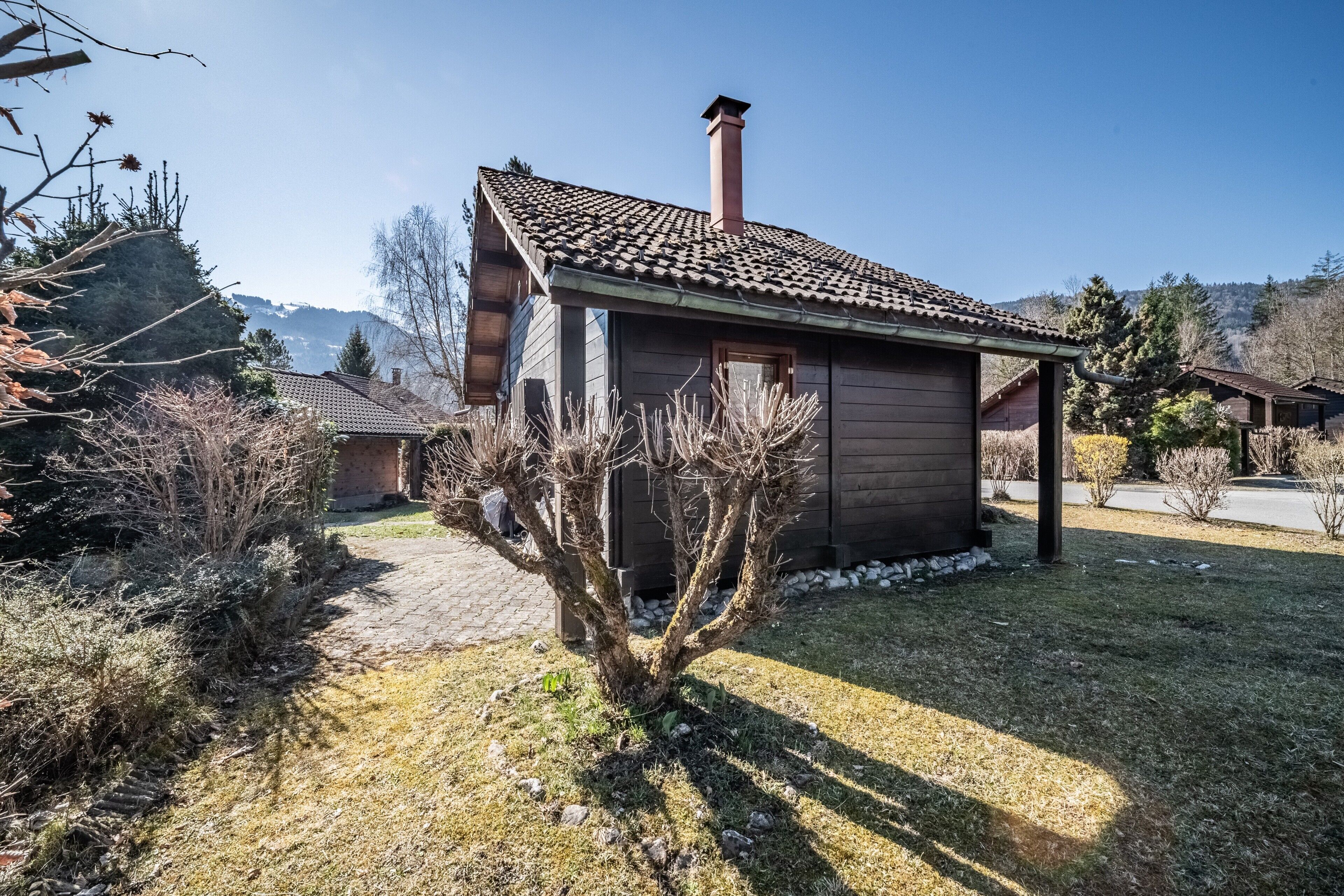 Une cabane en bois avec un toit en tuiles, entourée d’une zone herbeuse et d’une végétation clairsemée.