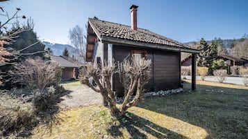 Une cabane en bois avec un toit en tuiles, entourée d’une zone herbeuse et d’une végétation clairsemée.