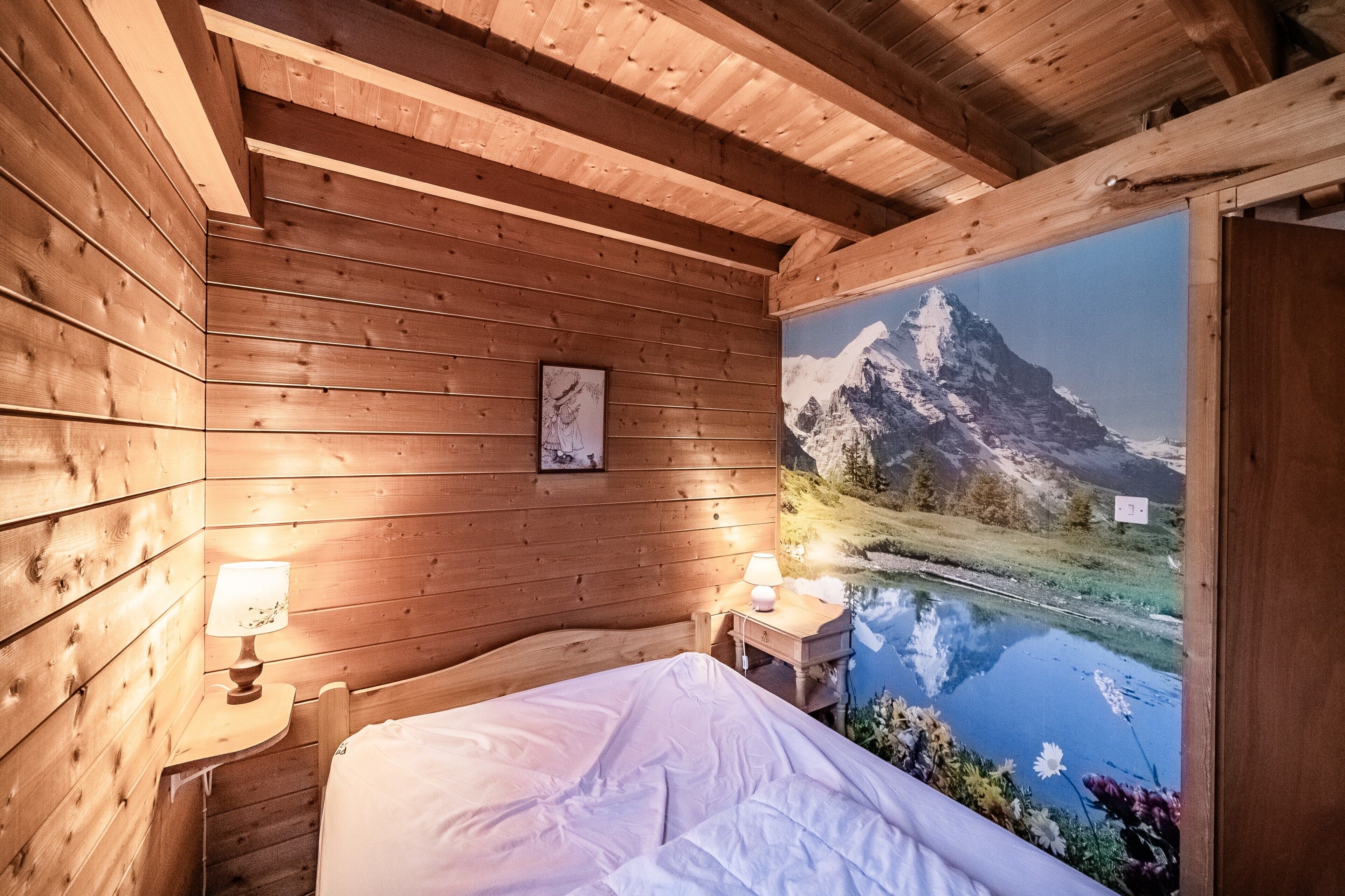Una habitación tipo cabaña de madera con una cama, una mesita de noche con lámpara y un gran mural en la pared con un paisaje montañoso.