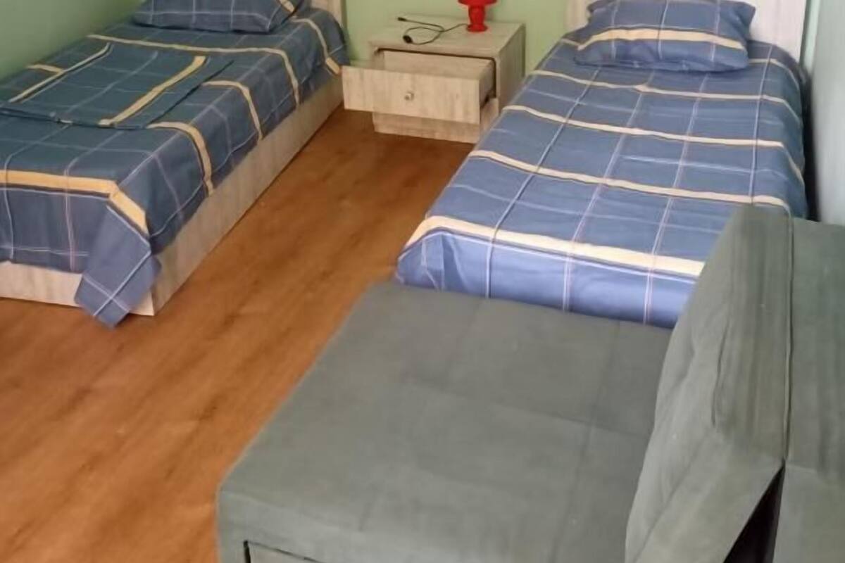 Ein Zimmer mit zwei Betten, jeweils mit einer blau-weiß karierten Tagesdecke, einem hölzernen Kopfteil und einem Nachttisch mit einer roten Lampe.