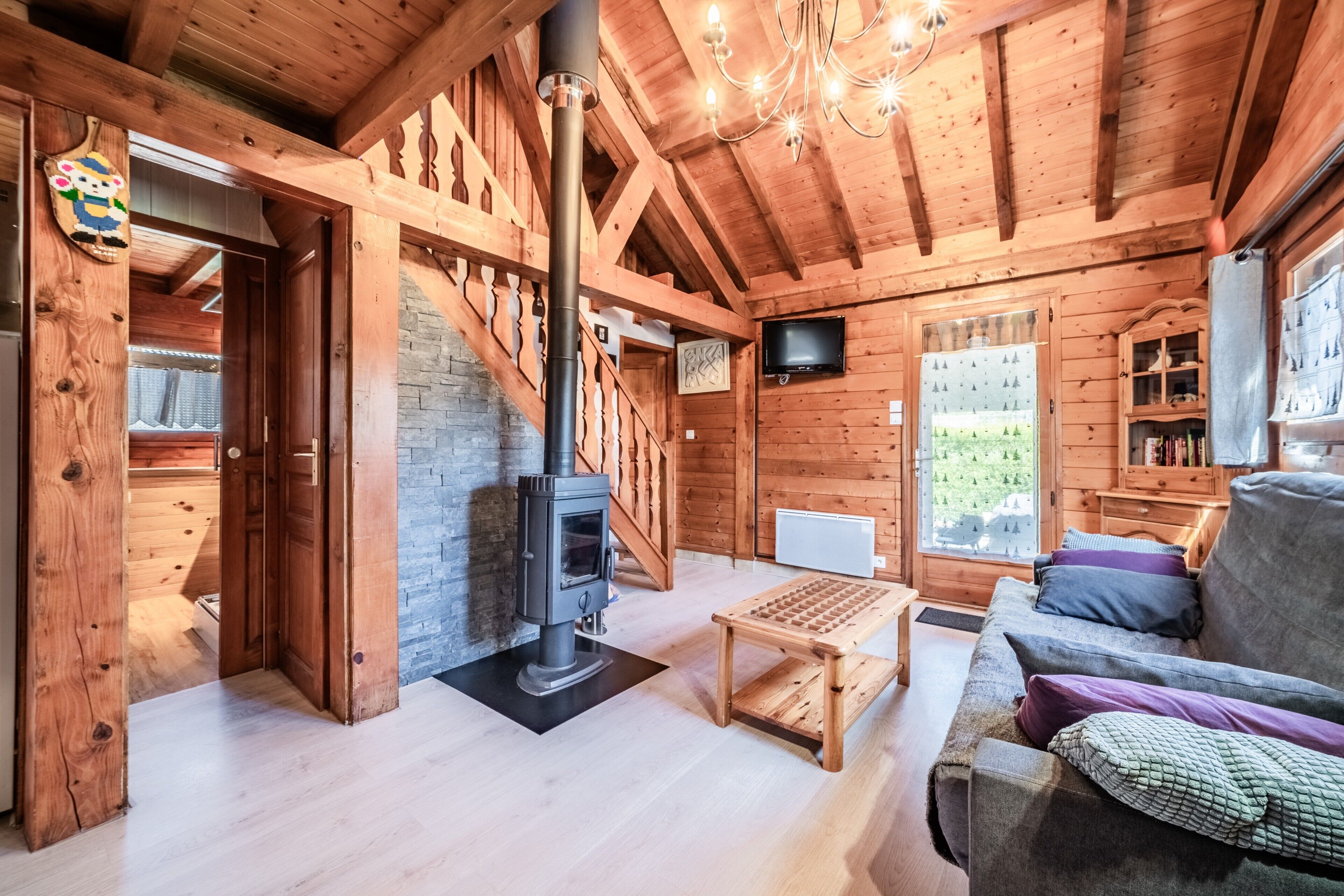 Interior de una cabaña de madera con chimenea, sofá, mesa y televisor montado en la pared.