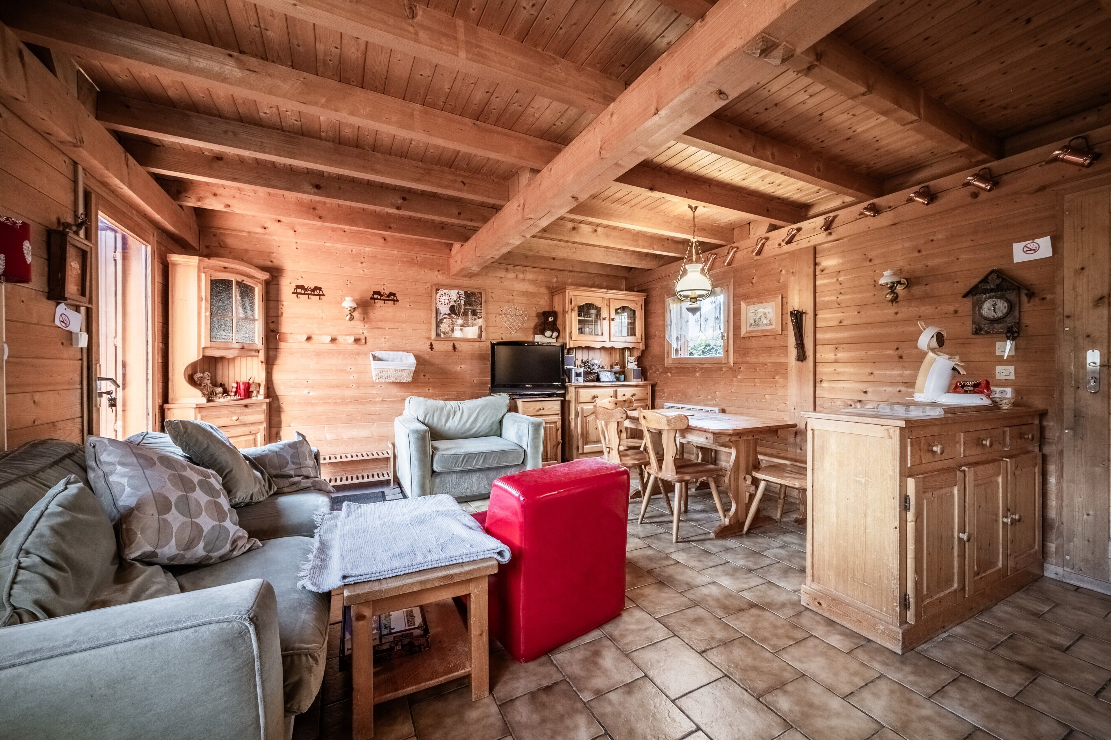Interior de una cabaña de madera con sofá, reposapiés rojo, mesa de comedor, zona de cocina y armarios de madera.