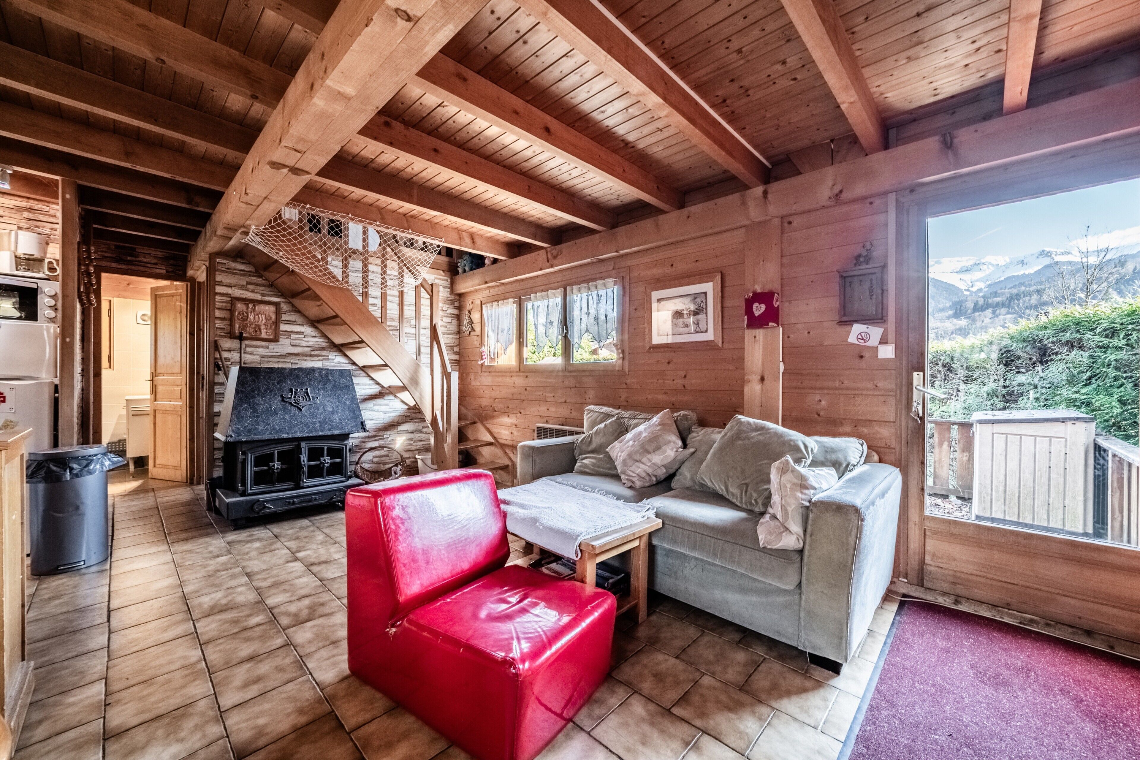 Interior de una cabaña de madera con chimenea, un sillón rojo, un sofá y vistas a la montaña.