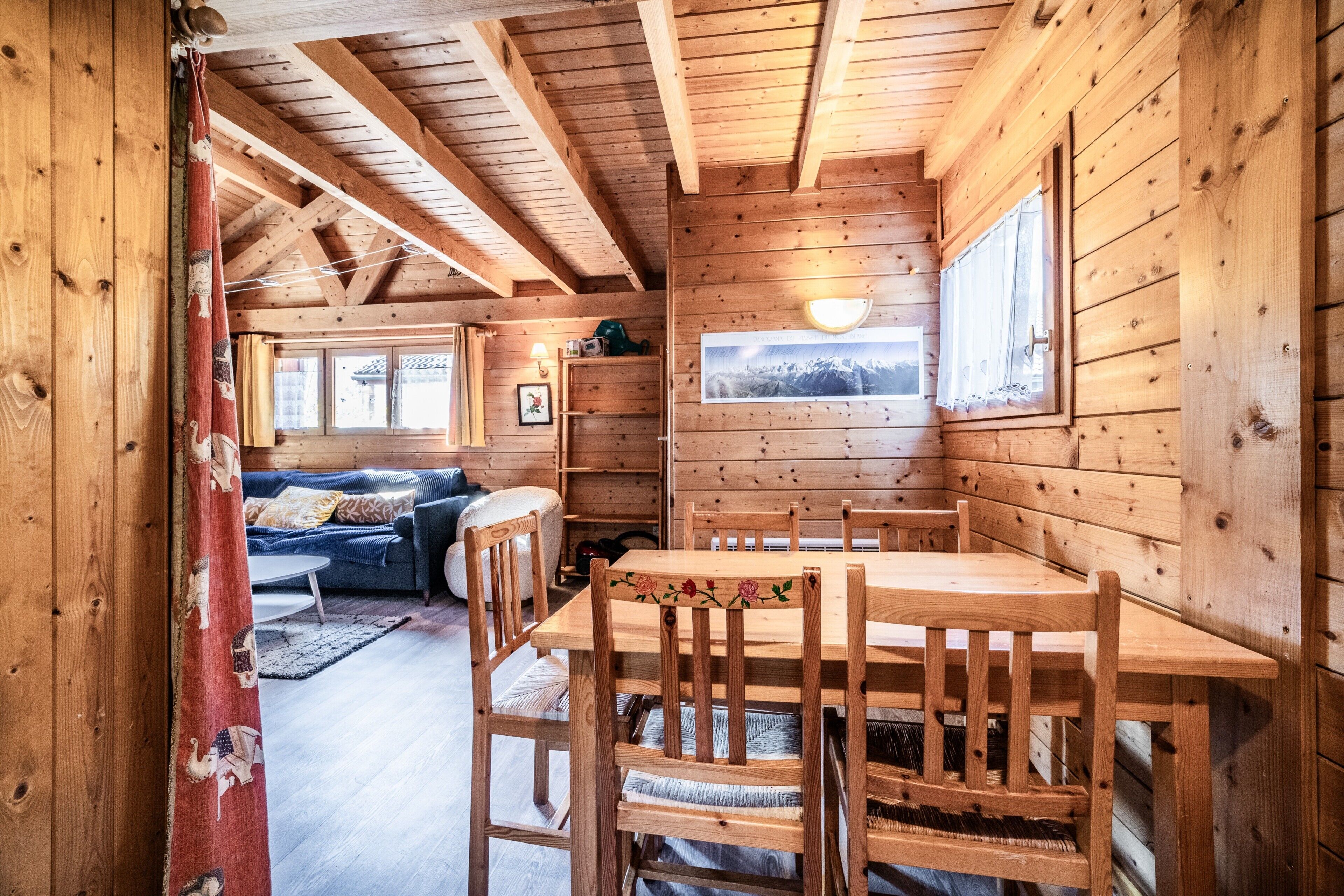 A wooden interior with a dining table, chairs, a sofa, and a window with a view.