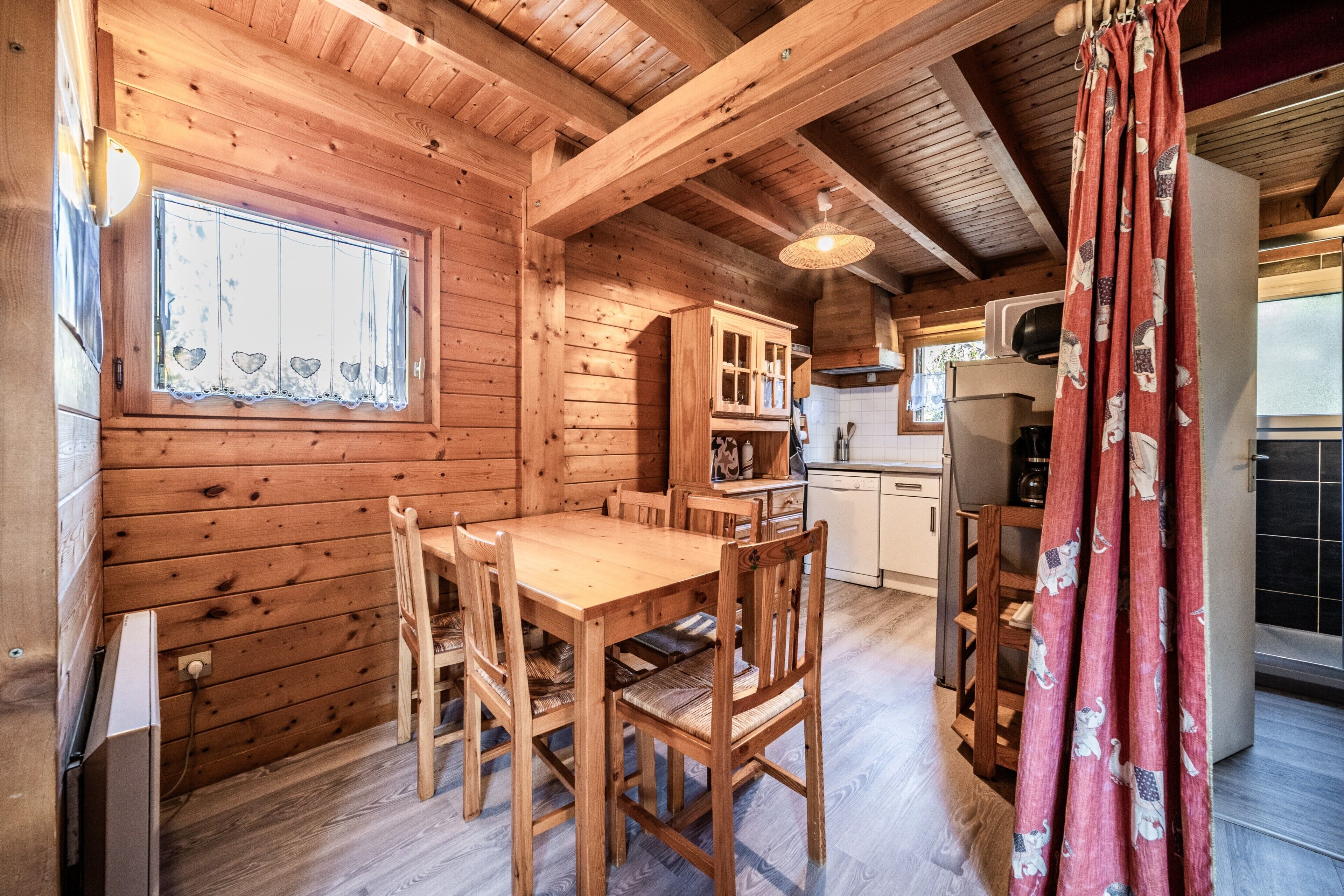 Un intérieur en bois comprenant une table à manger, des chaises, un coin cuisine et une fenêtre agrémentée d’un rideau décoratif.