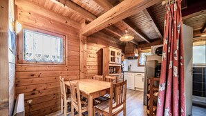Un intérieur en bois comprenant une table à manger, des chaises, un coin cuisine et une fenêtre agrémentée d’un rideau décoratif.