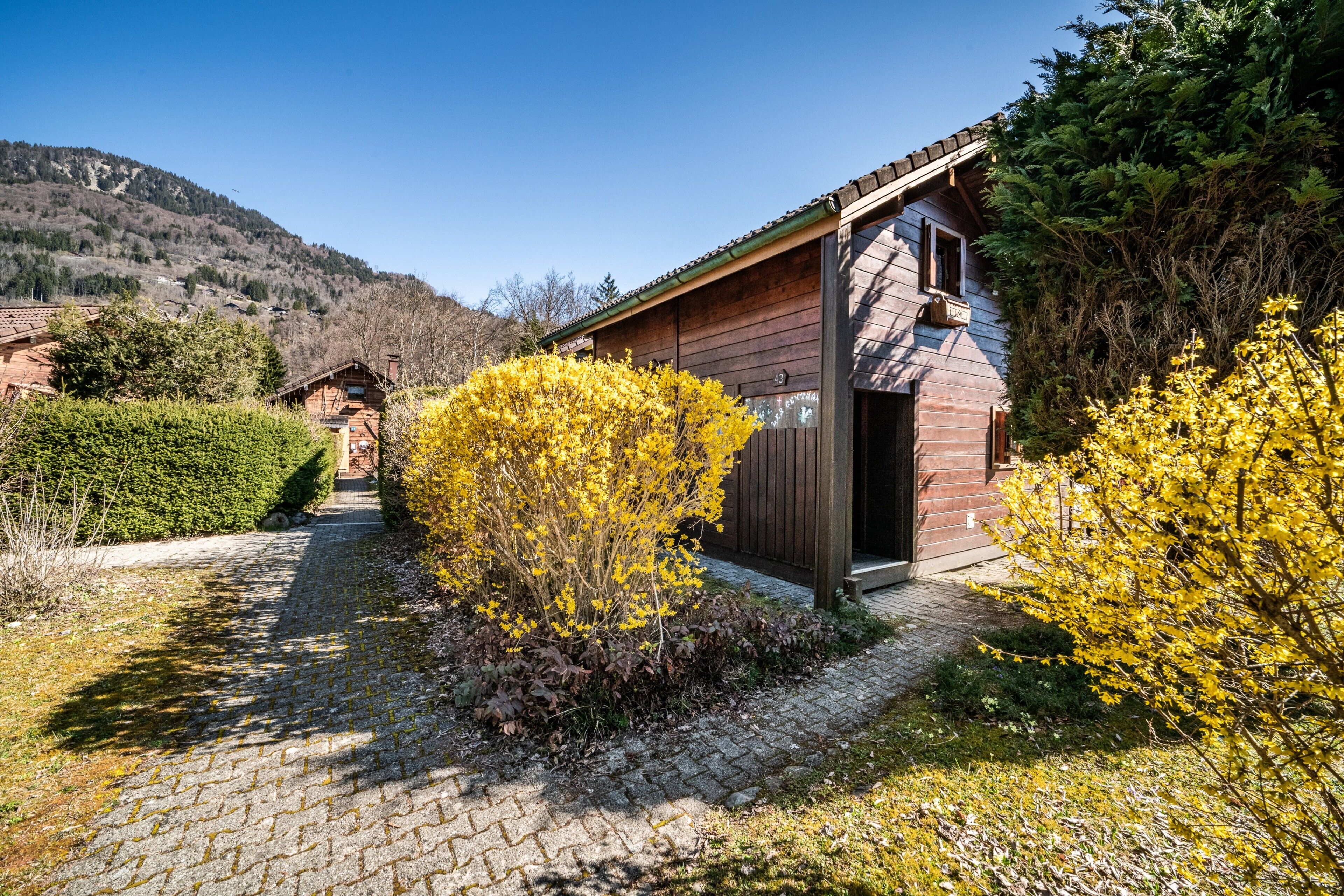 Eine Holzhütte mit Backsteinfassade, umgeben von einem Kopfsteinpflasterweg und gelb blühenden Sträuchern.