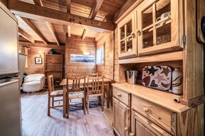A wooden cabin interior with a dining area, kitchen, and a window with a mountain view. - Chalet “6p Classic - Les Gentianes” with Mountain View, Private Terrace & Wi-Fi (Verchaix)