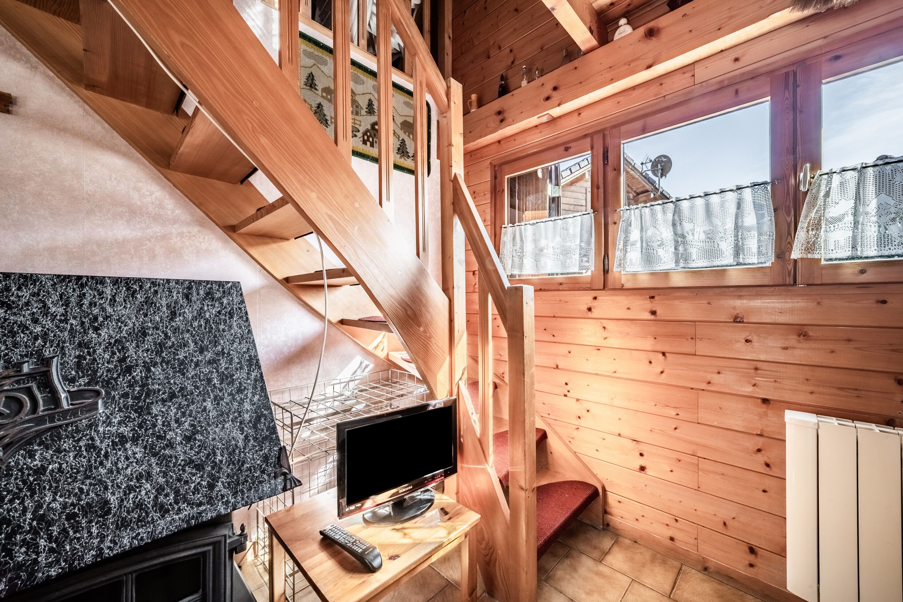 L’intérieur d’un chalet en bois, avec un escalier en bois, un téléviseur posé sur un meuble en bois et une fenêtre avec un rideau.