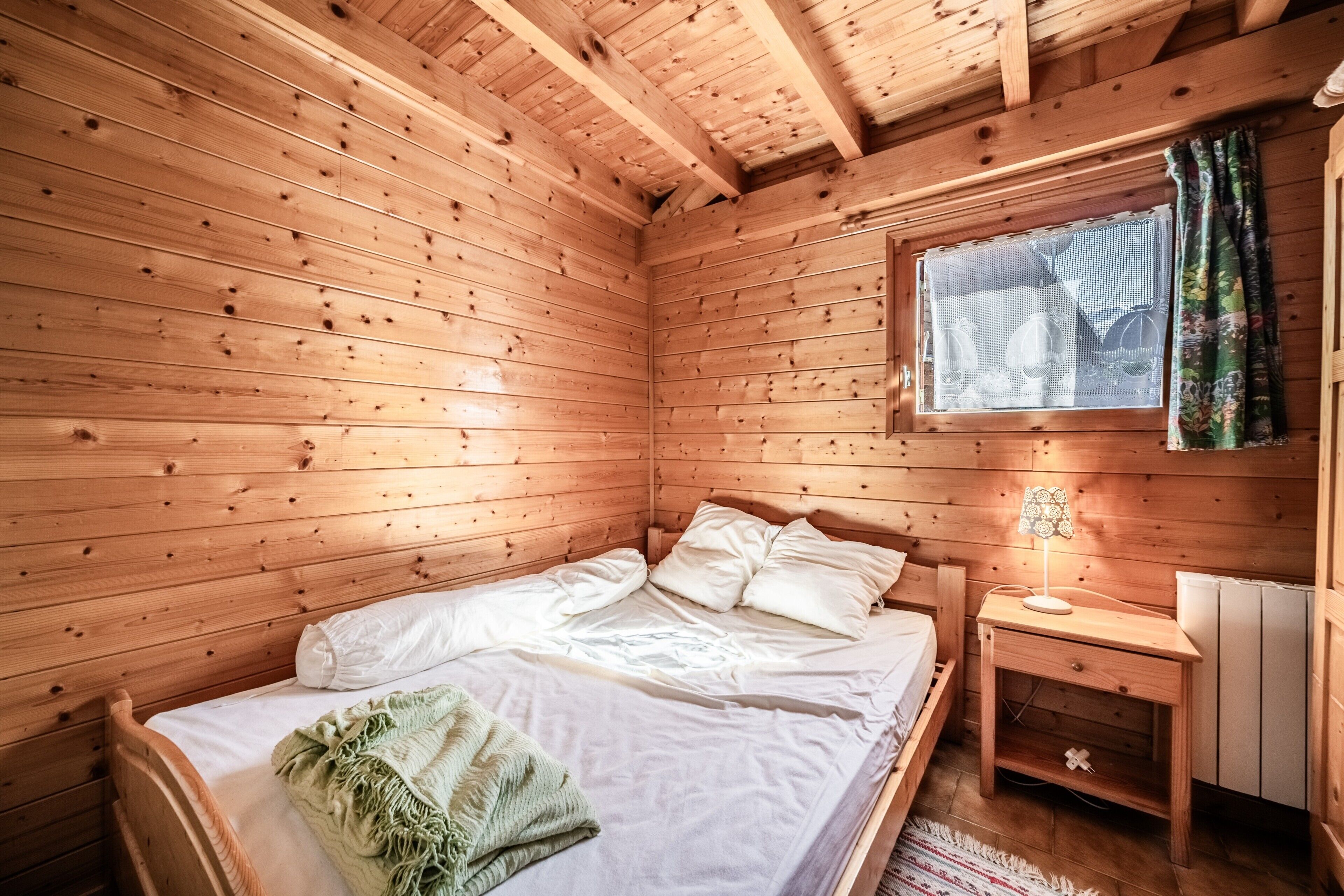 Una camera in una baita di legno, con un letto, un comodino, una finestra con tende e un termosifone.