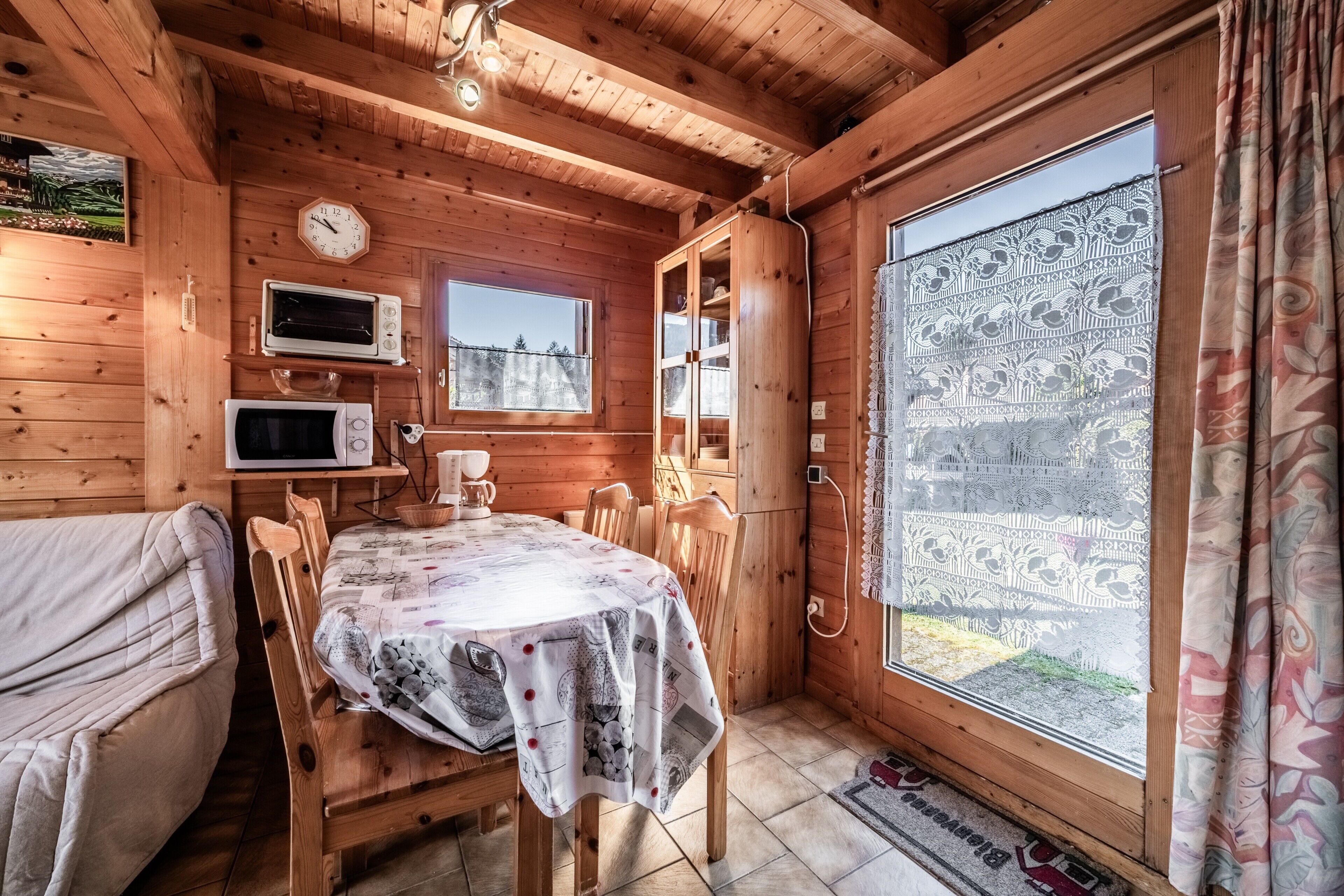 Interior de una cabaña de madera con una mesa de comedor, sillas, un reloj y una ventana con cortinas.