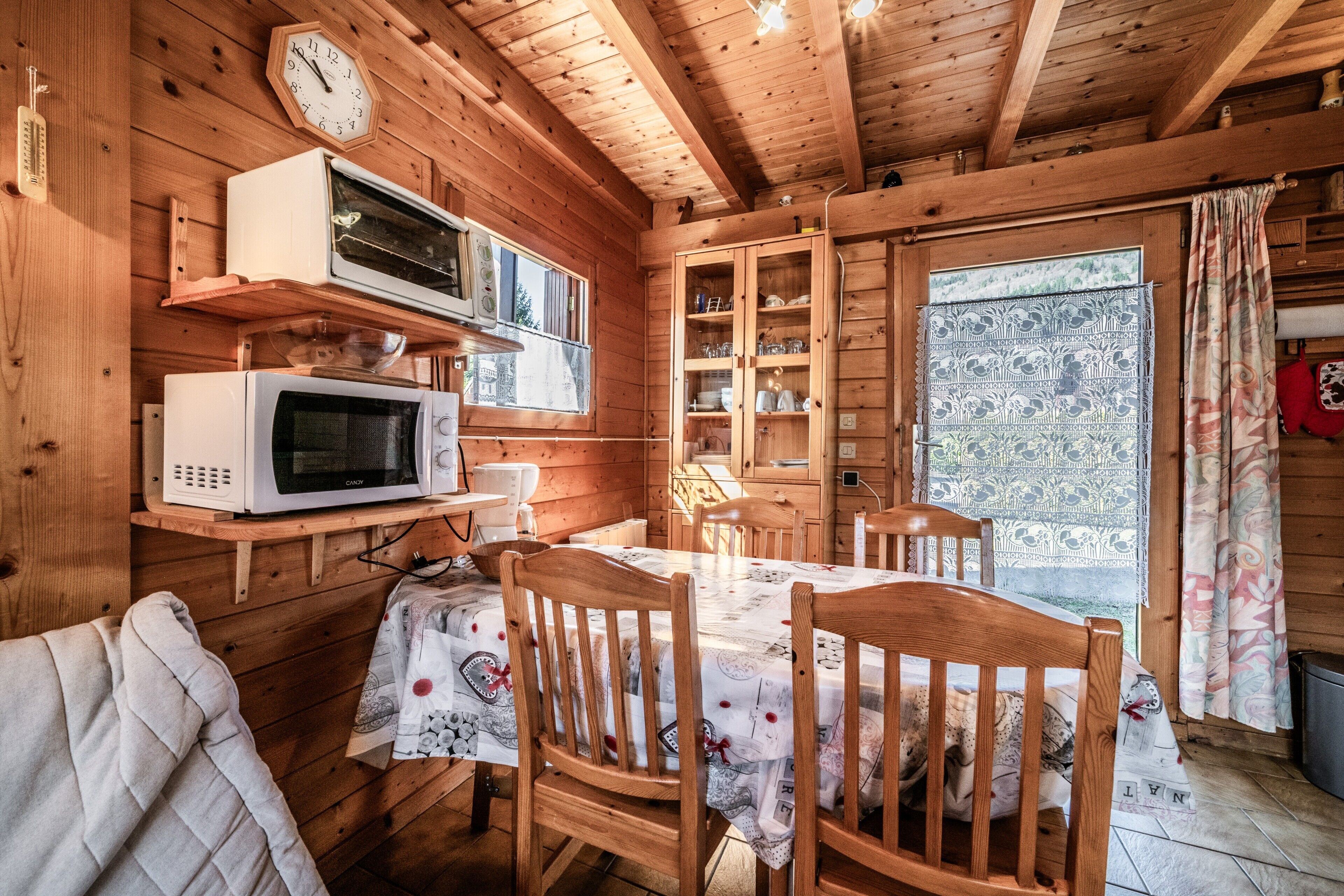 Interno di una baita in legno con un tavolo da pranzo, sedie, un forno a microonde, una sveglia e una finestra con tende.