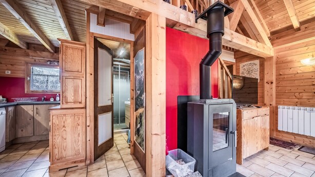 Houten cabin interieur met een keuken, een rode accentmuur, een houtkachel en een zichtbare deur naar een andere kamer.