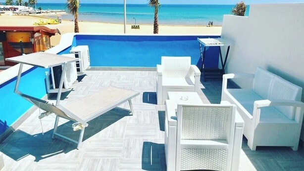 A rooftop pool area with white lounge chairs and a table, overlooking a beach with palm trees and a clear blue sky.