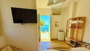 A bedroom with a bed, a TV, a wicker chair, a wooden wardrobe, and a view of the sea through an open door.