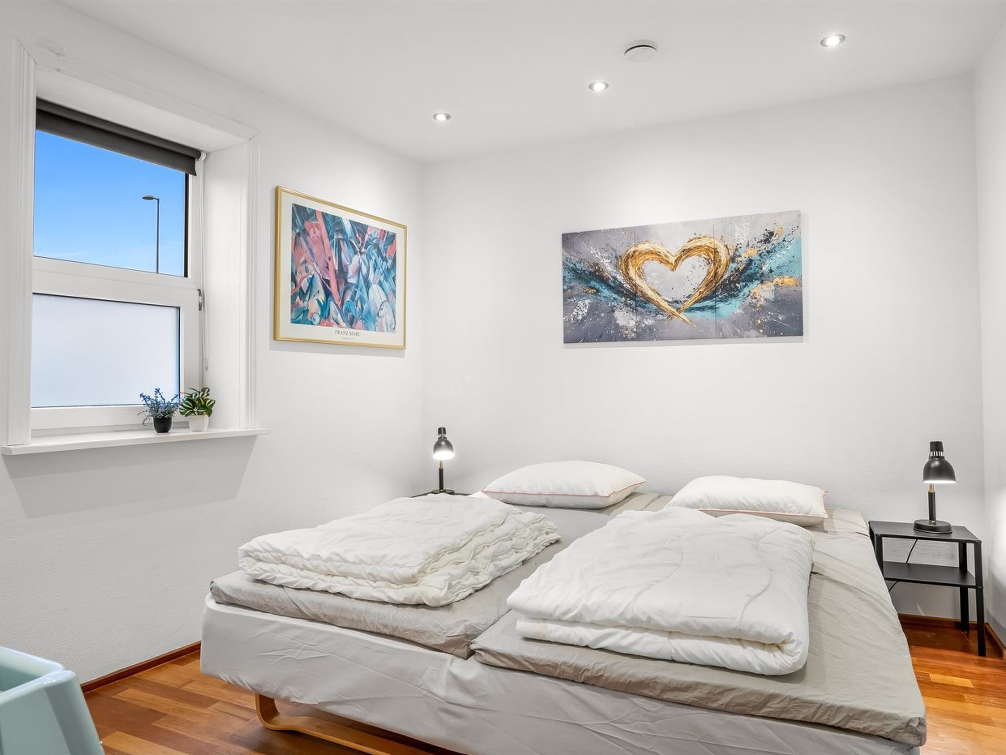 A bedroom with a bed, two bedside tables, a window, and two paintings on the wall.