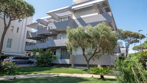 Un immeuble moderne à plusieurs étages, doté de balcons, avec une voiture garée devant et un arbre au premier plan.
